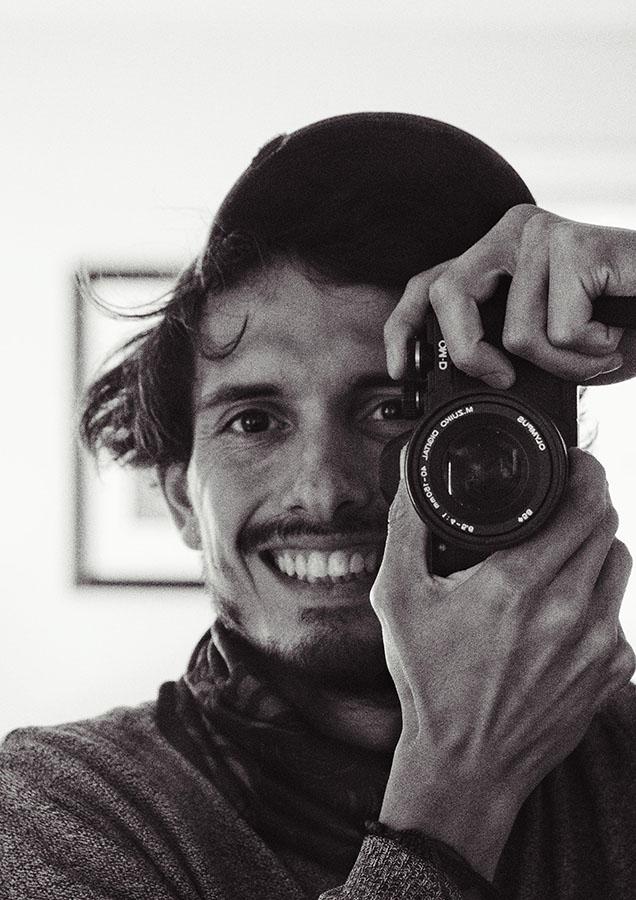 A black and white photo of a person holding a camera up near their face to take a photo while smiling