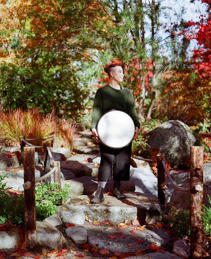 A person in a green sweater standing on stone steps in a garden and holding a drum