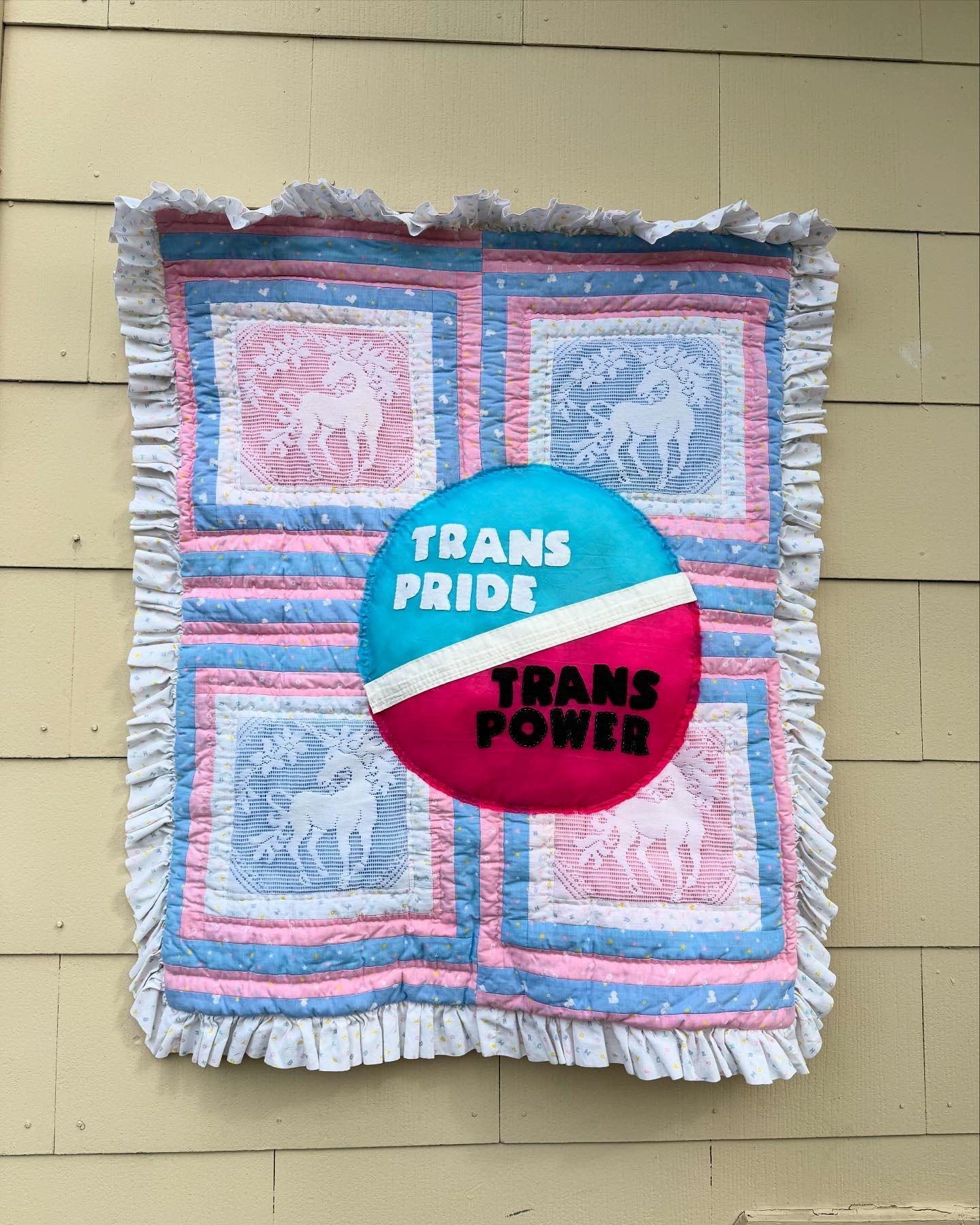 Photo of a blue, pink, and white quilt with unicorns and a bright circle containing the words "TRANS PRIDE / TRANS POWER"