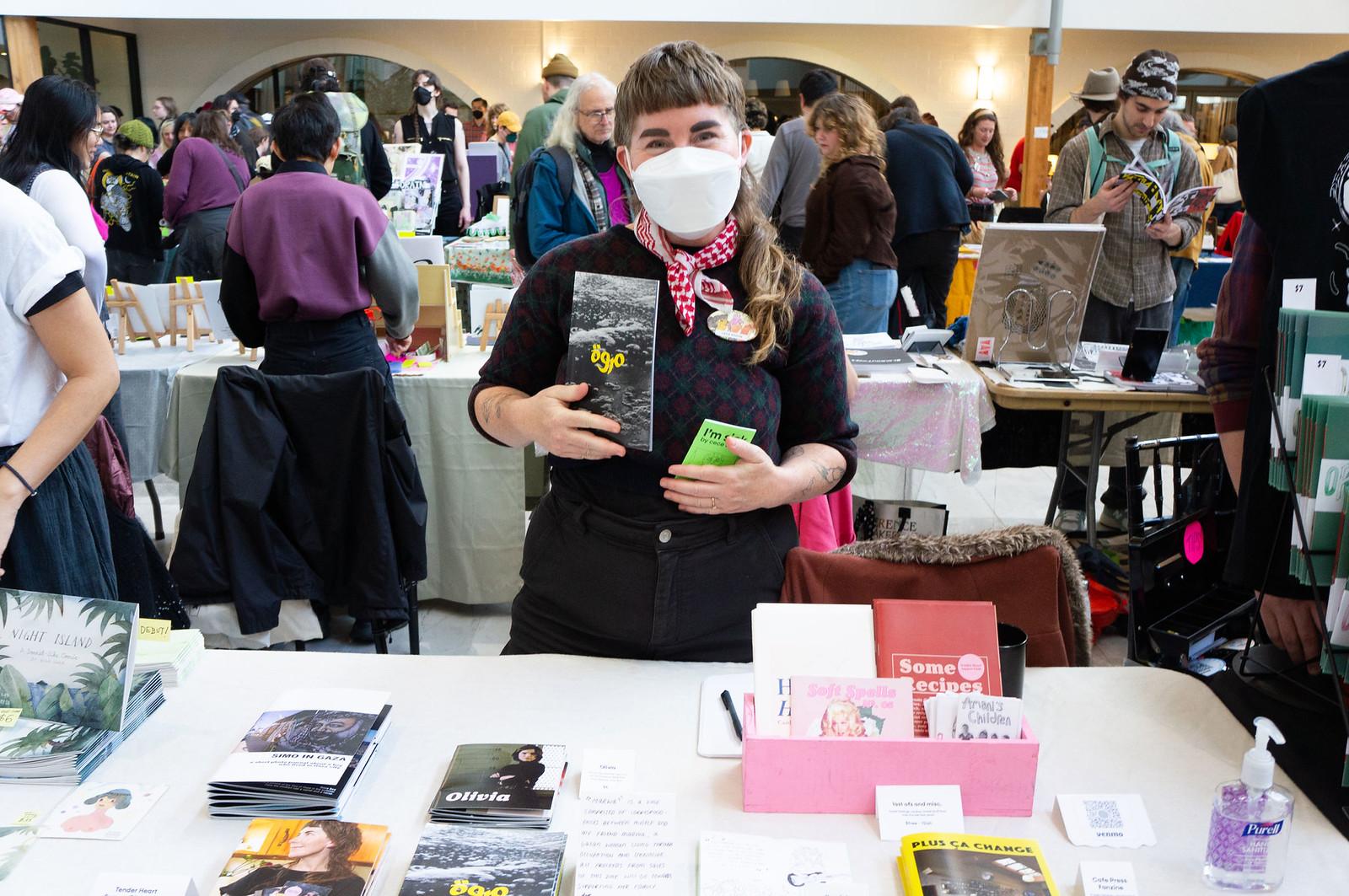 Cece Edson standing behind a table laid with zines holding example zines up