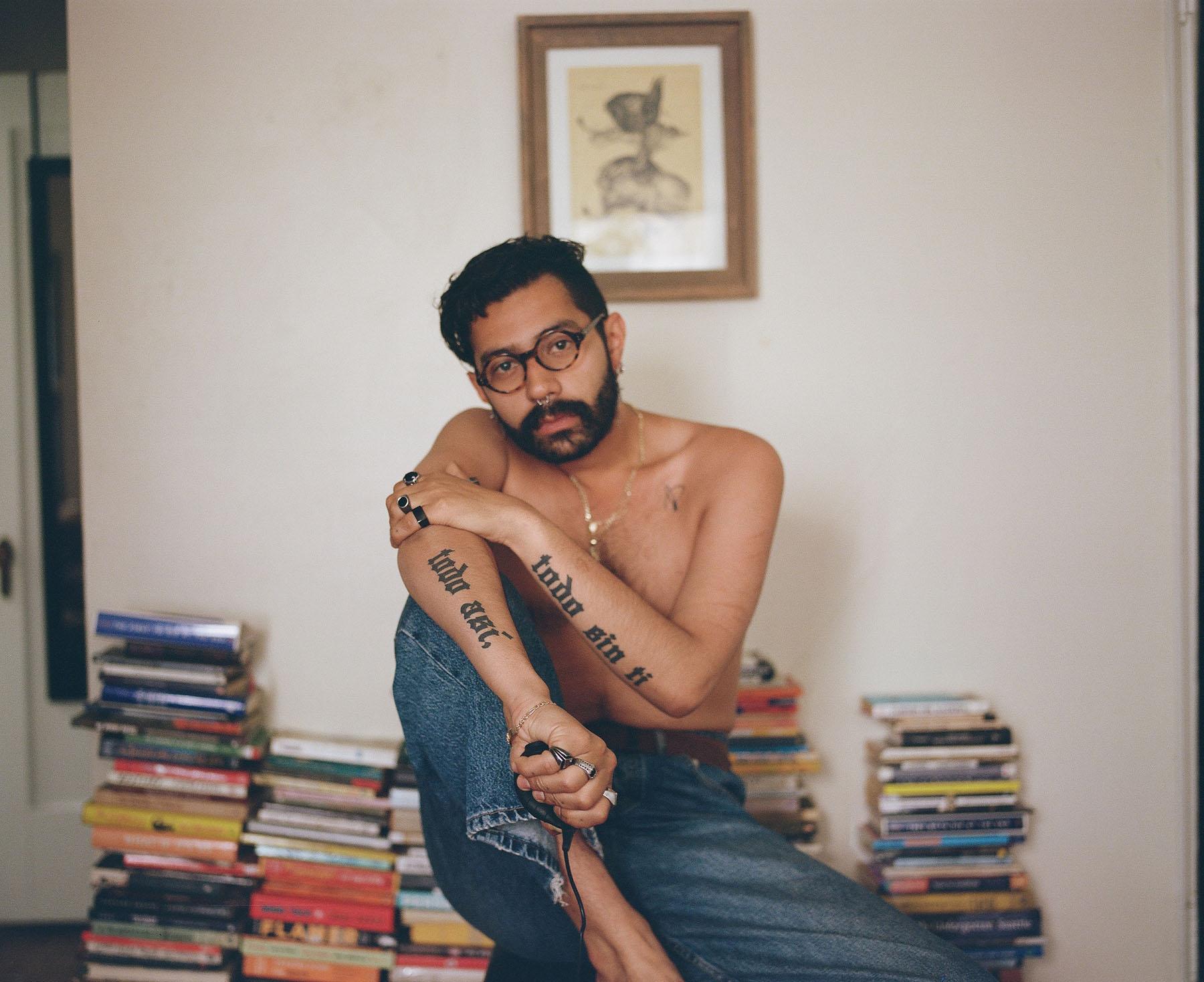 A portrait photo of a person in jeans and no shirt sitting on a desk with piles of books behind them