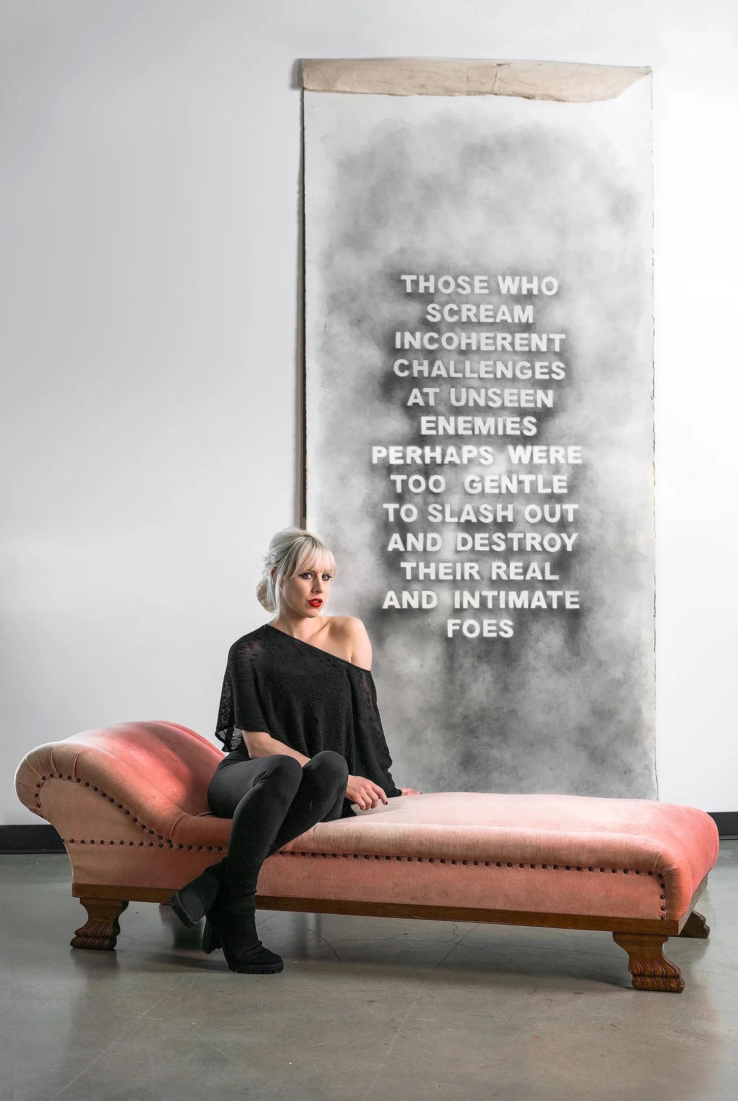 Woman with short blonde and black dress hair sitting on a red/orange chaise lounge chair with an artwork handing on the wall behind her