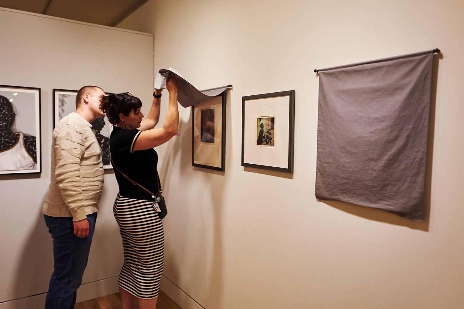 A couple of people looking at an artwork that one of them is lifting a curtain to see