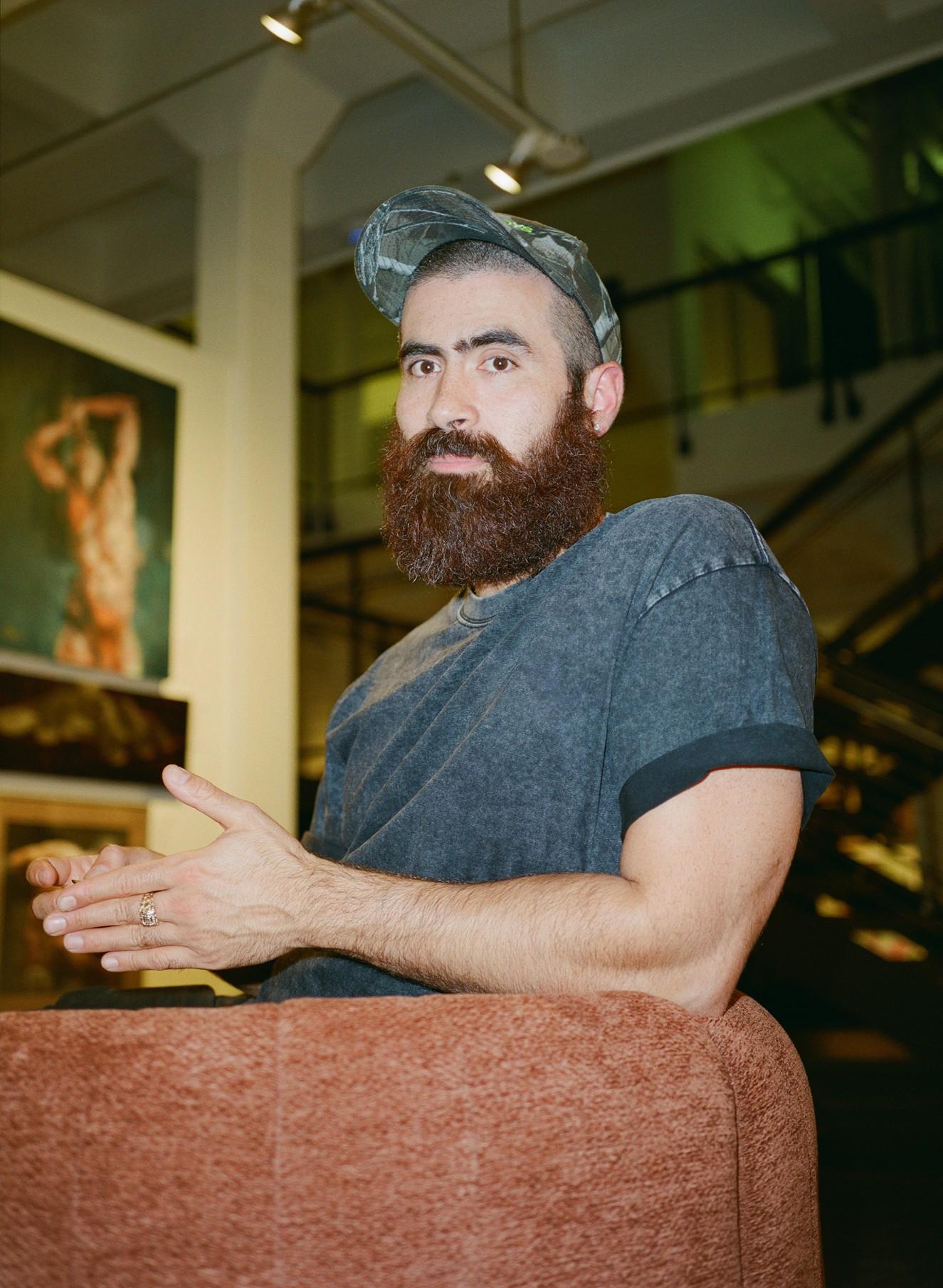 A man with a large beard wearing a cap seating on a couch or furniture