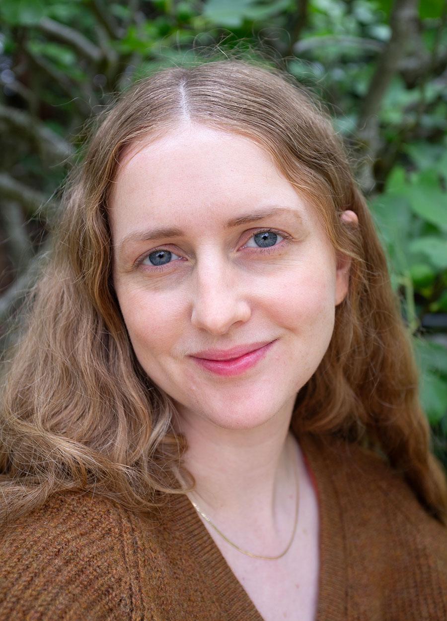 A tightly cropped portrait photo of a slightly smiling woman outdoors with greenery behind her