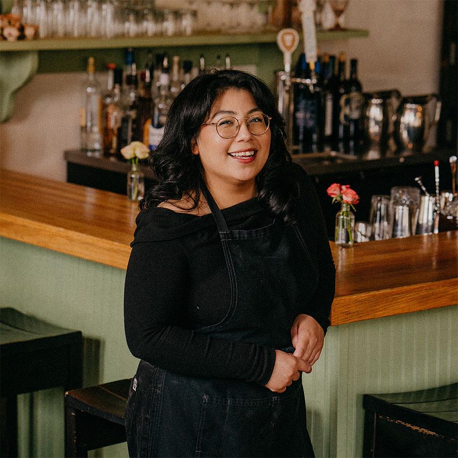 A woman in a black top and dark apron leaning on a corner of a bar and smiling