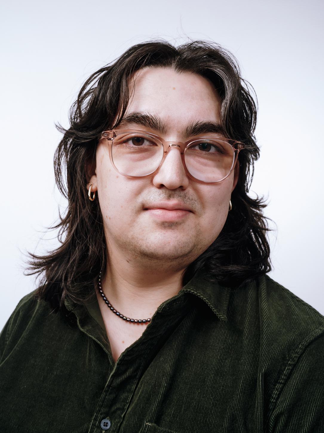Headshot of Zack Kunisawa-Conklin, they have shoulder length dark brown hair, and are wearing clear-rimmed glasses and an olive corduroy button up shirt
