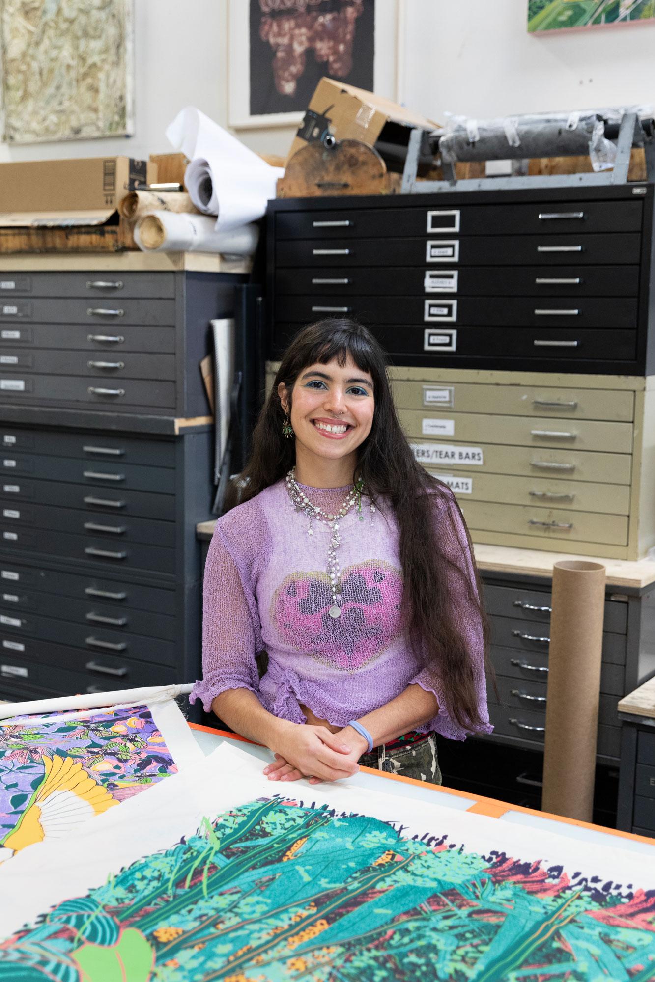 Artist María Zamora smiles at the camera with one of her screenprints laid out on the table in front of her
