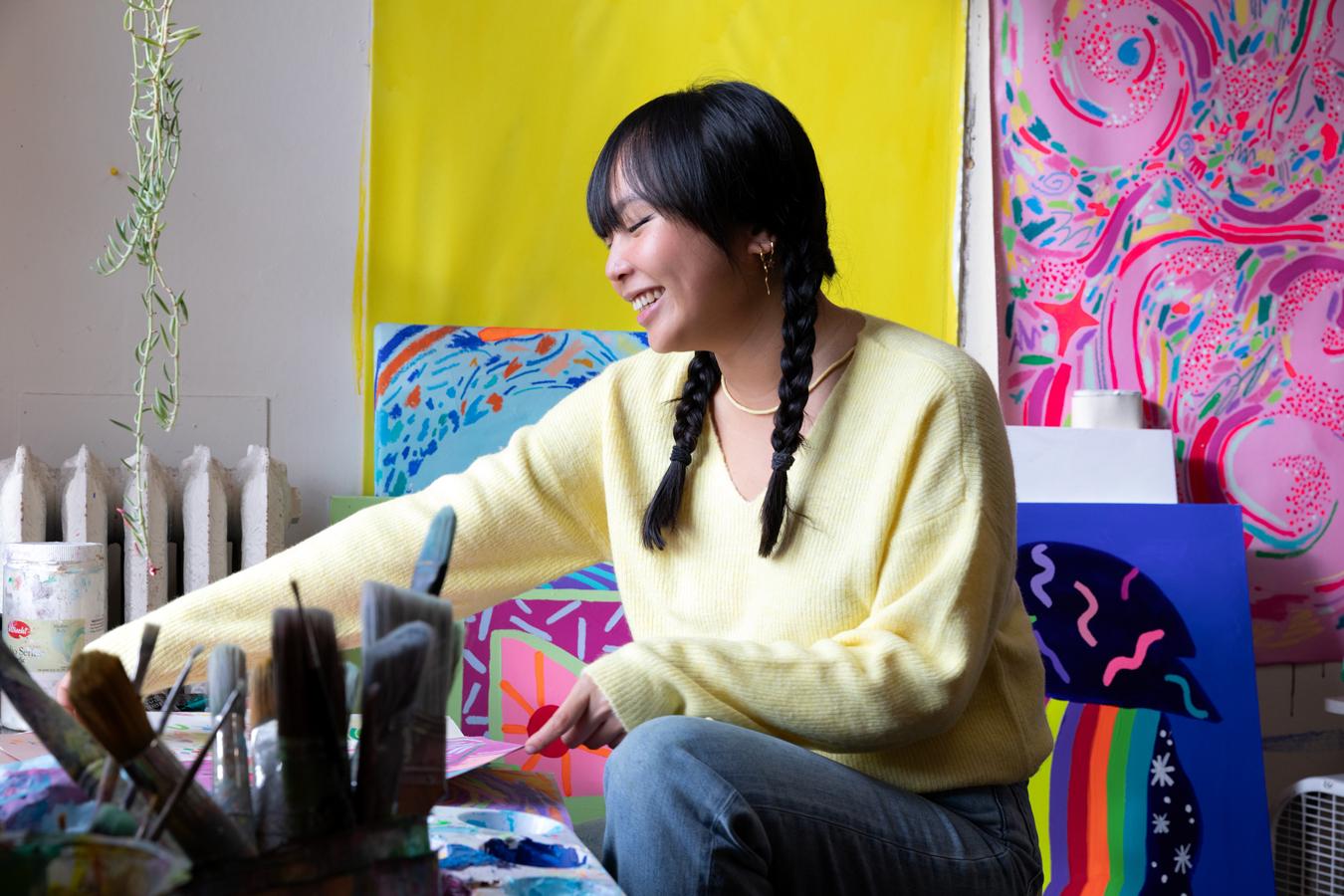 Photo of Nikita Ares, a woman with dark hair, smiling and looking at her art works on a table and surrounded by her bright art