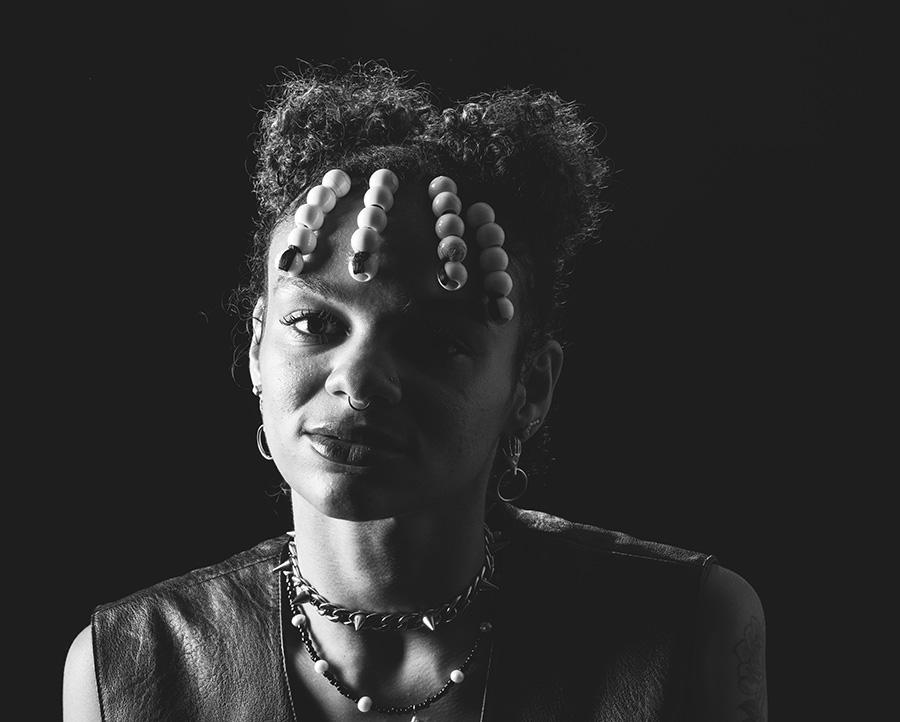 A black and white portrait photo of a woman with beads in her hair and a number of piercings and pieces of jewelry