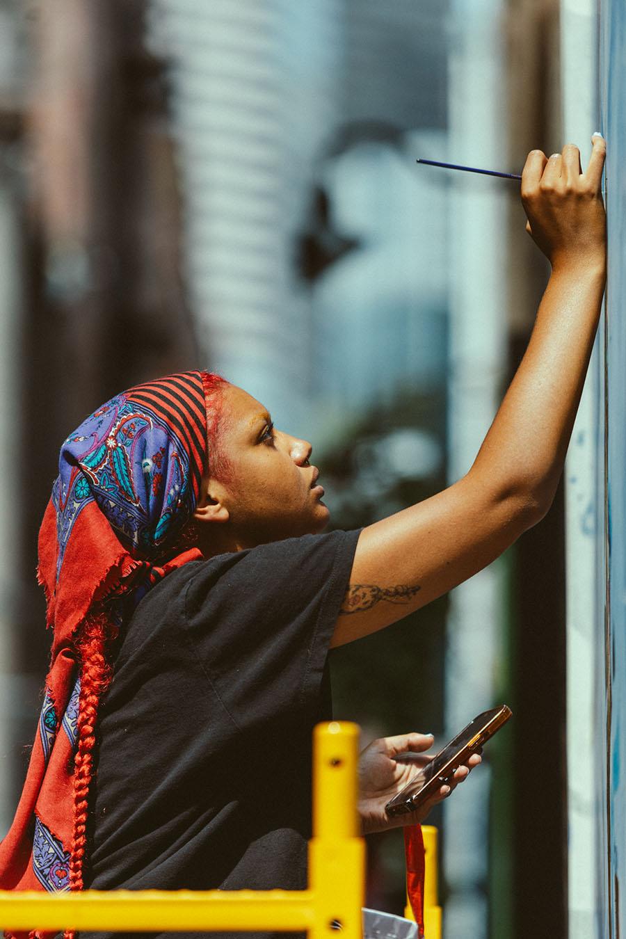 A person in profile reaching up and painting on a large wall or mural
