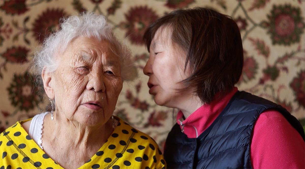 A middle age person whispering something into an older person's ear in front of a wall covered in wallpaper