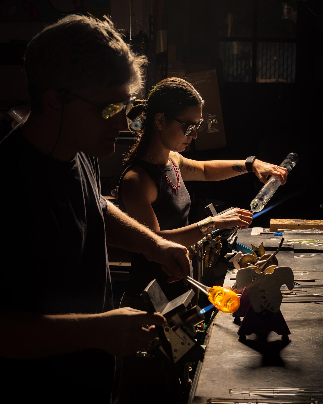 Two people working on glass works standing side by side at a work table while wearing safety glasses
