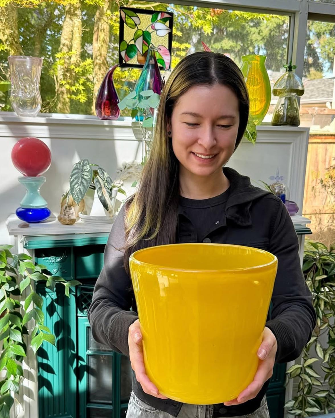 A woman with long dark hair with dyed yellow parts smiling and looking down at a large yellow glass vessel in her hands