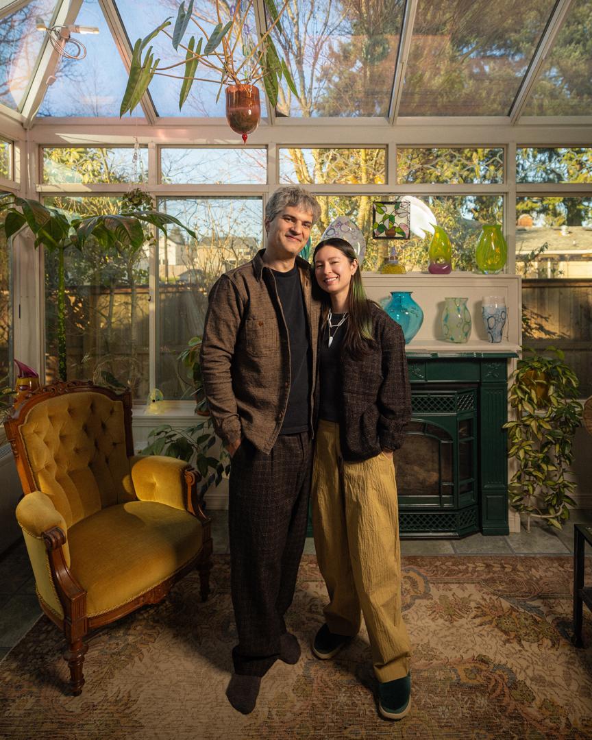 A couple, a man and a woman, standing a well lit sun room next to a yellow armchair