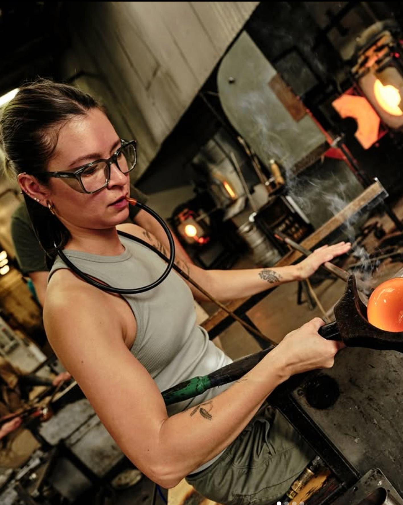 Sue Kim working on a molten piece of glass in a hot shop wearing safety glasses and a gray tank top.