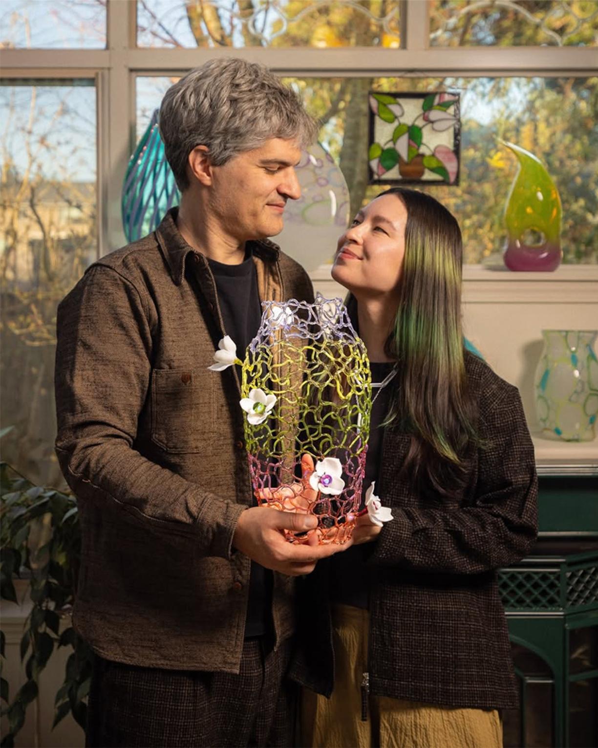 Zeke Spier and Sue Kim standing a well lit sunroom smiling at each other while holding a delicate glass sculpture