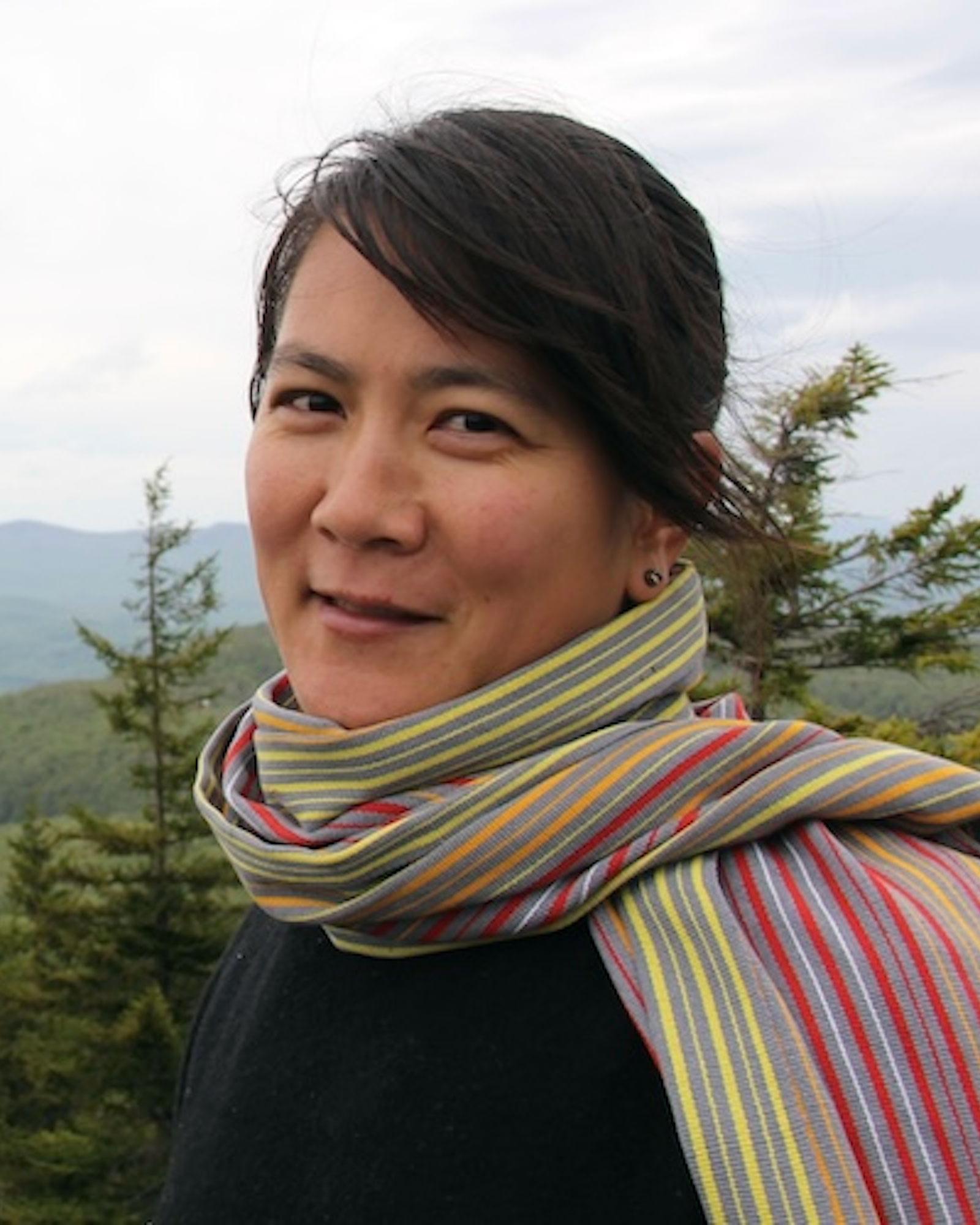 Yoshi Nakagawa, a woman with dark brown hair pulled back wearing a stripped green, white, and pink scarf and a black top