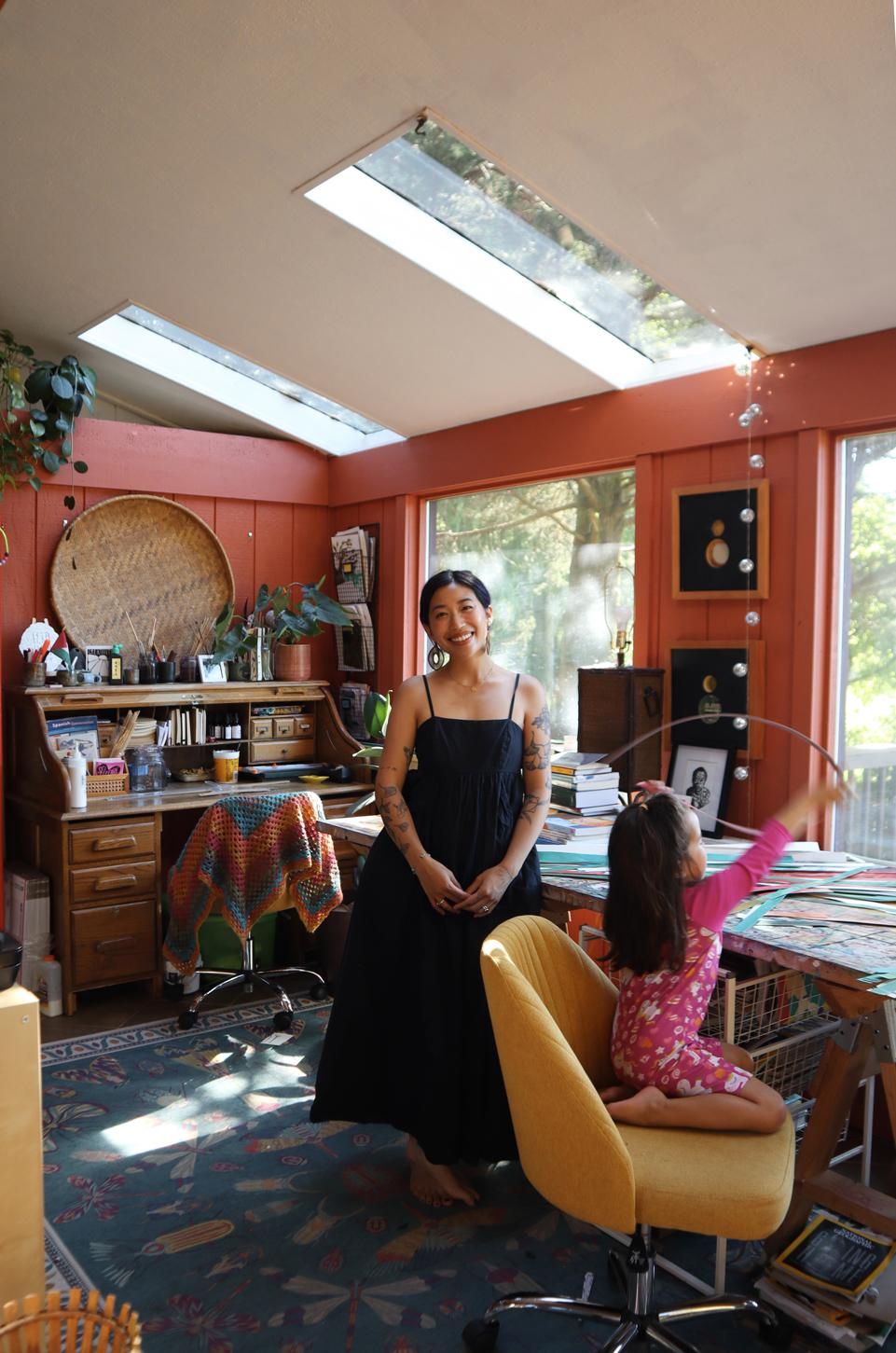 lexa toledo villanueva stands smiling in a black dress in her studio while a young child plays in a chair by the work table