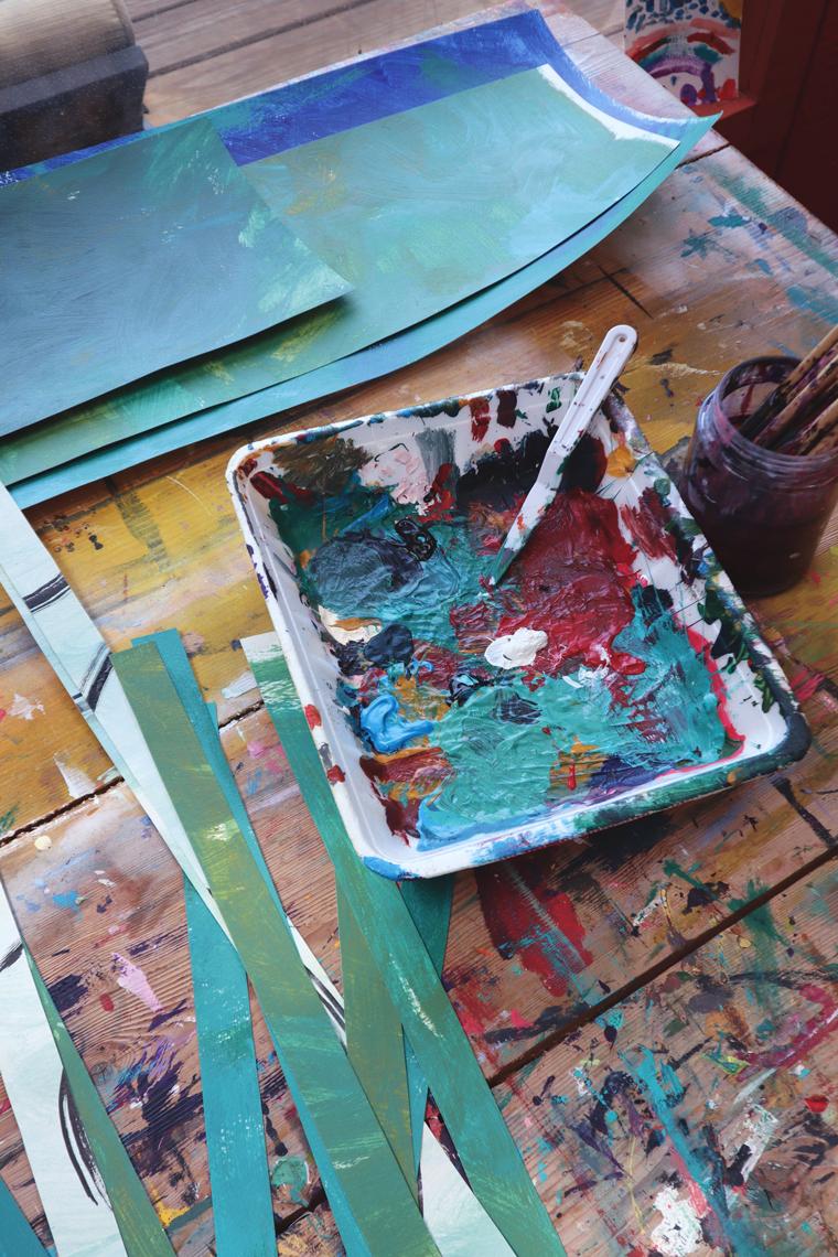 A tray with dried out paint on a work table beside stripe of colorful painted paper and paintbrushes in a jar