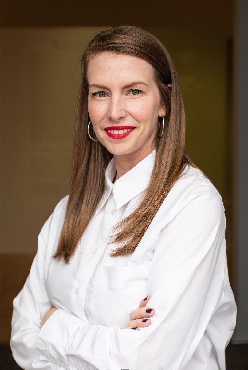 A professional headshot style photo of a smiling woman with brown hair wearing a white blouse