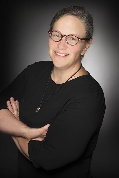 Photograph of Keri Pollock, a woman with short hair and red glasses wearing a black shirt