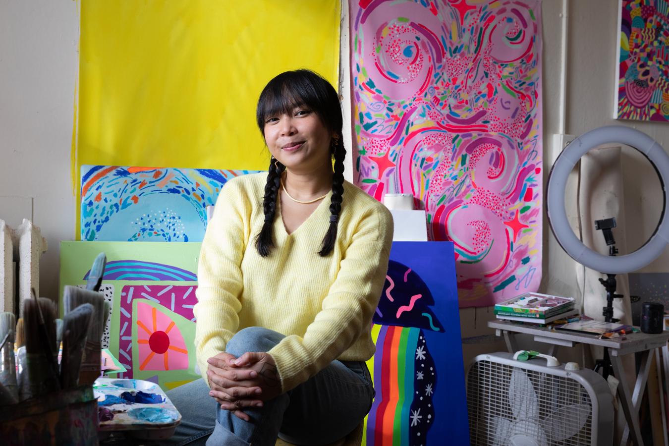 Photo of Nikita Ares, a woman with dark hair, smiling surrounded by her colorful paintings