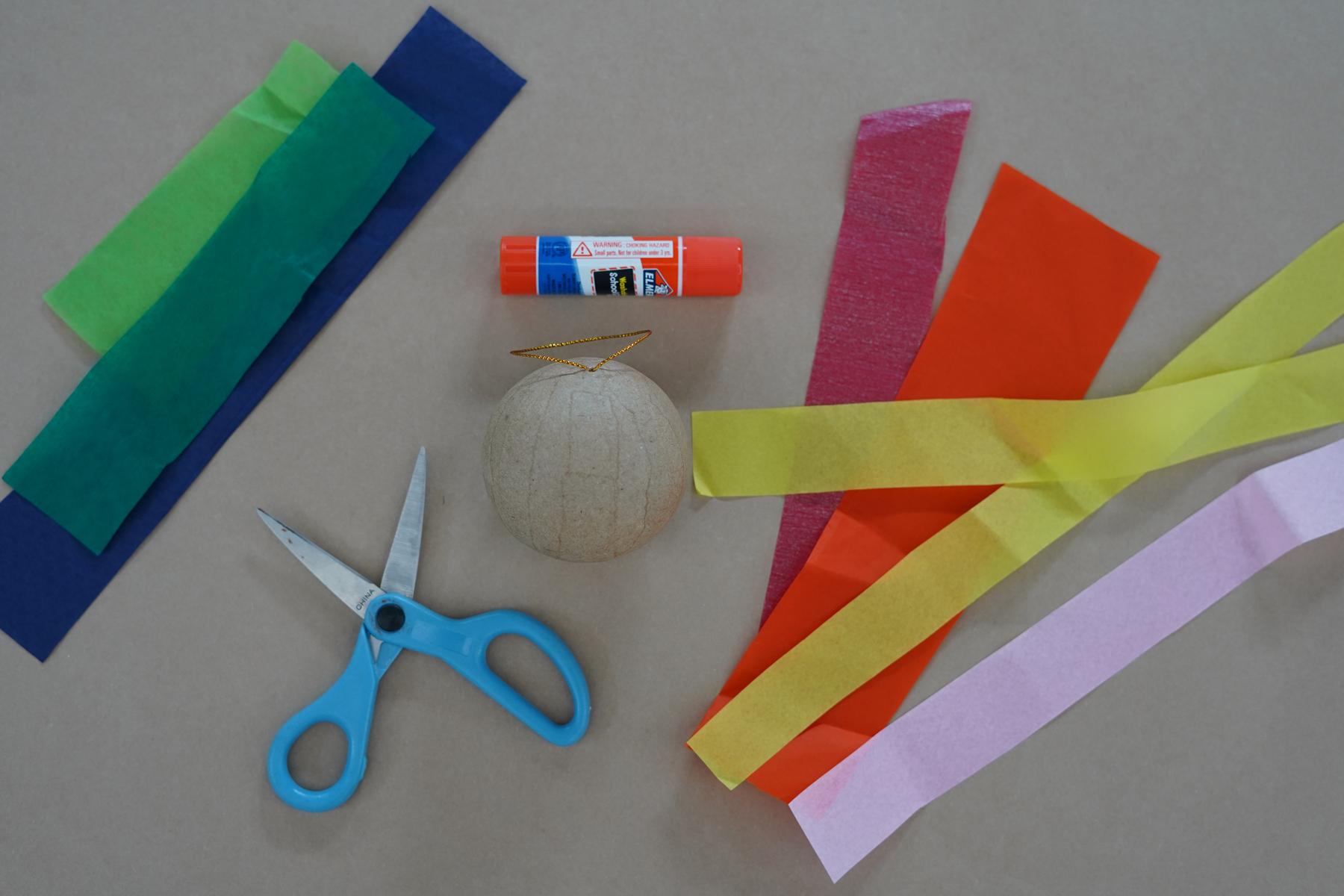Photo of art supplies on a table including tissue paper in a variety of colors, scissors, a glue stick, and a cardboard ball