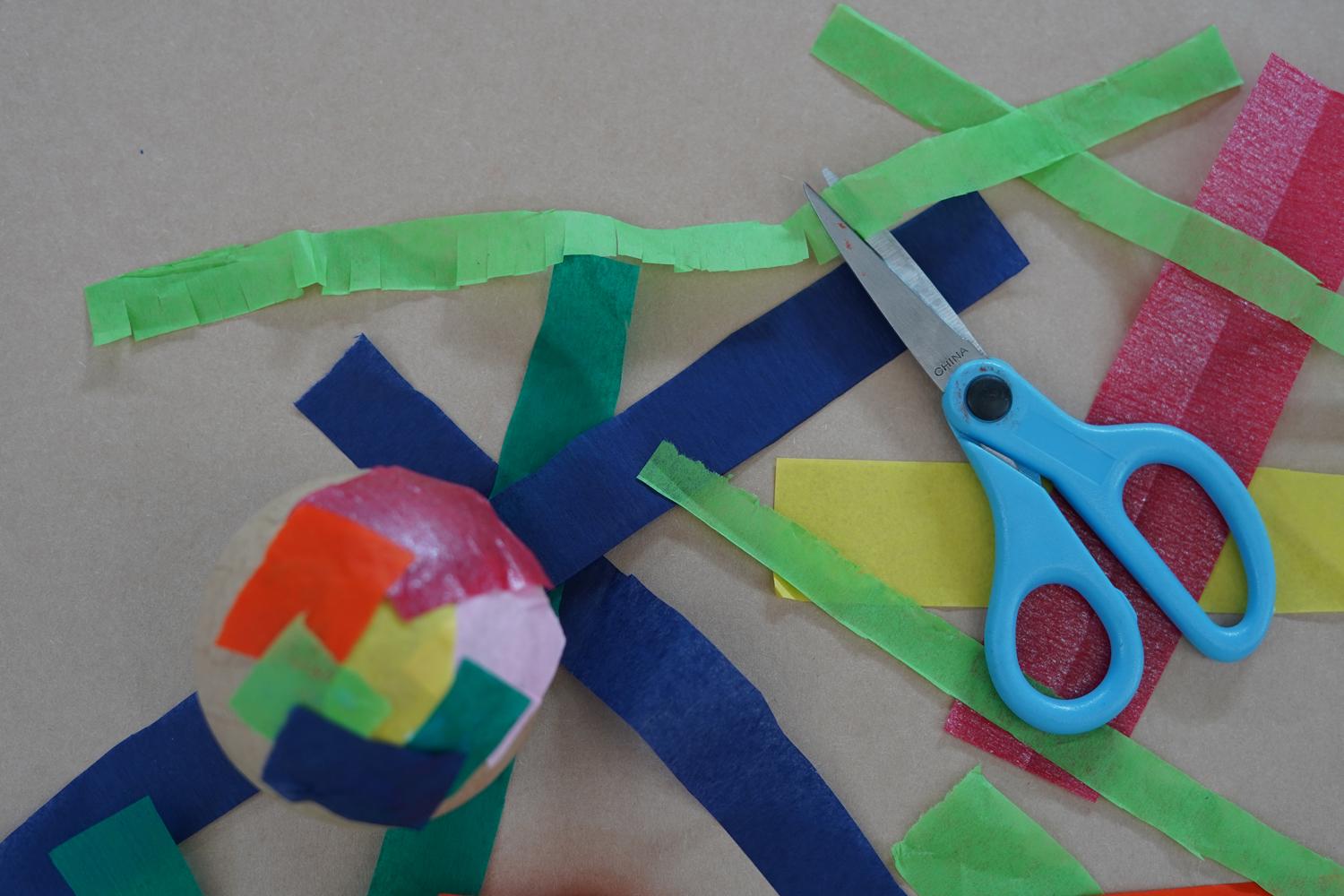 Photo of a cardboard ball with pieces of tissue paper on it and a long piece of tissue paper with cuts into it making fringe on a tabletop