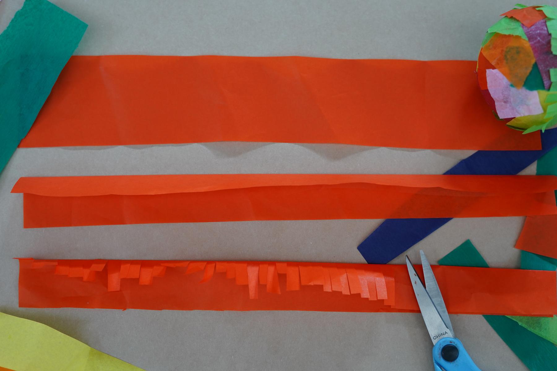Photo of three pieces of tissue paper: one wide flat piece, one piece folded in half, and the last piece folded with cuts in it making a fringe effect