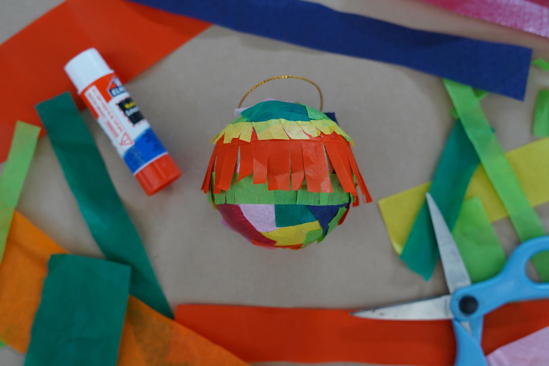 Photo of a cardboard ball covered with tissue paper fringe, surrounded by other tissue paper, a glue stick, and a pair of scissors