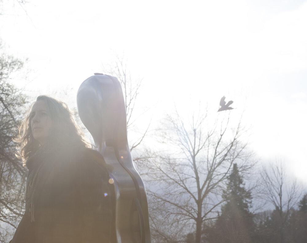 A woman in front of trees carrying a cello case on her back. A bird is flying by in the background.