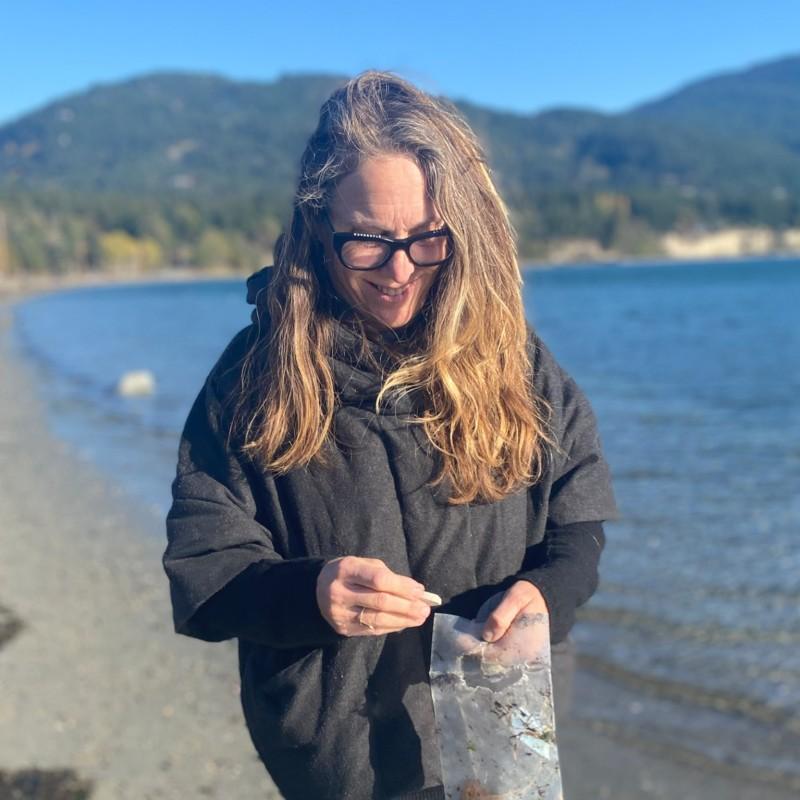 Woman with long blonde hair in a grey jacket standing on the beach with a plastic bag in her hand