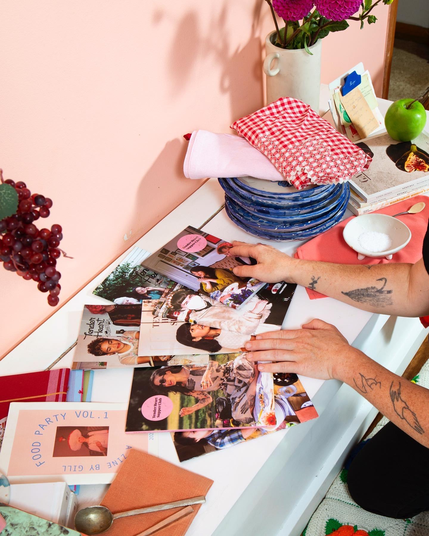 A bunch of zines on a narrow table with someone's hands sorting through them