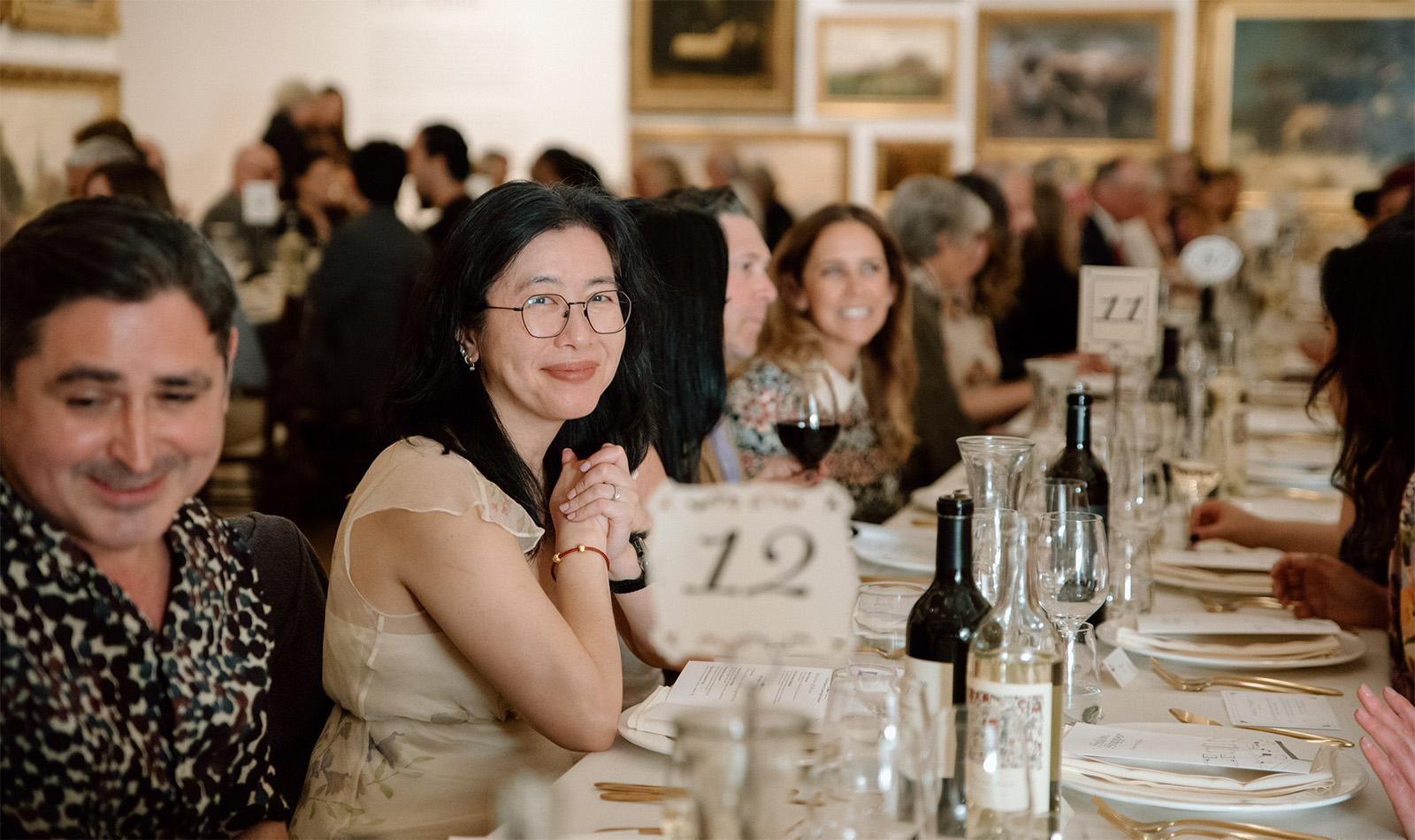 A woman surrounded by people smiling and sitting at a long table laid for dinner in an art gallery