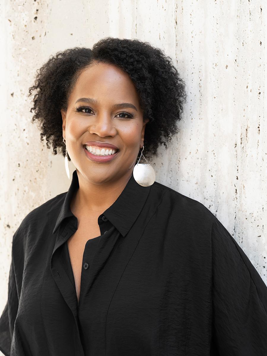 A smiling woman in a dark shirt standing against a wall