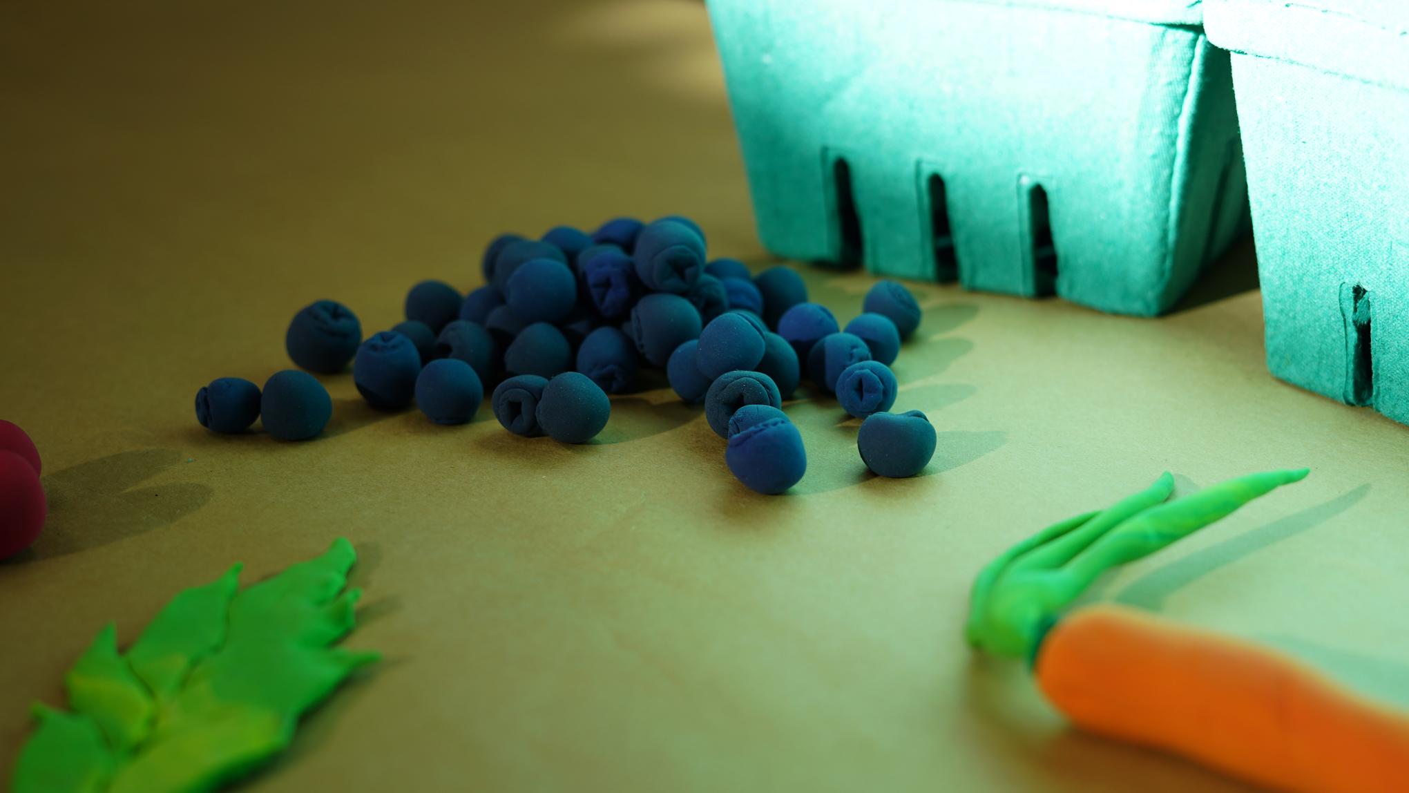 Photo of blueberries, lettuce, and a carrot made from modeling clay on a surface next to some green molded pulp berry baskets