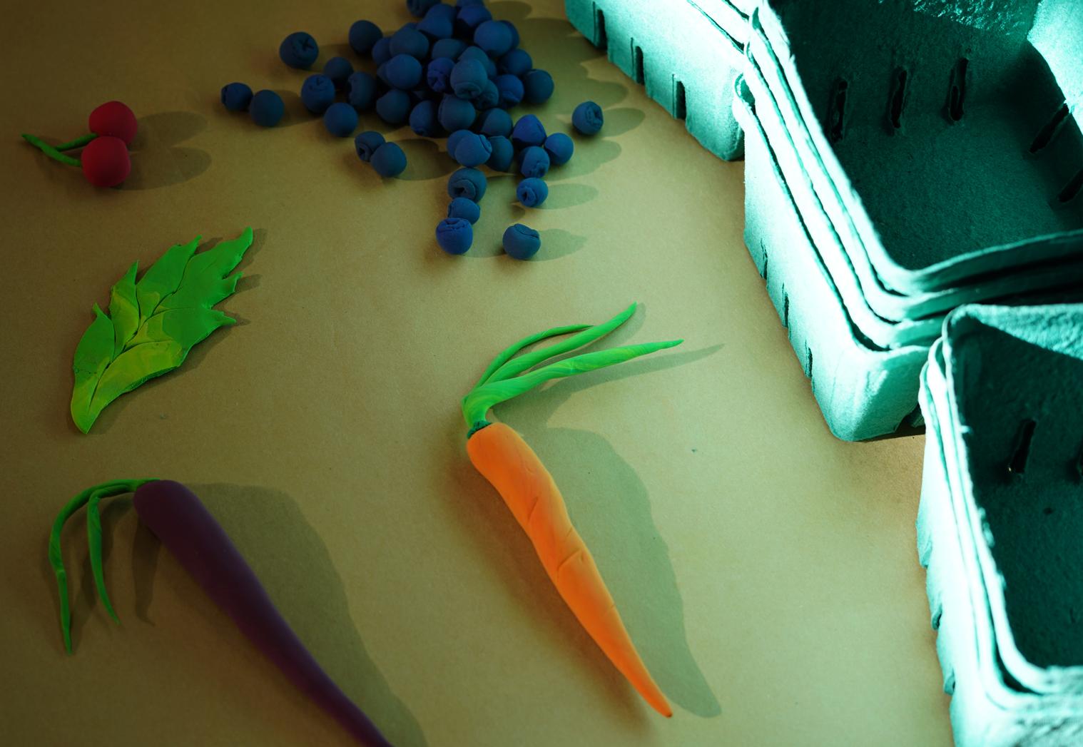 Photo of fruits and vegetables made of modeling clay on a table beside green molded pulp berry baskets