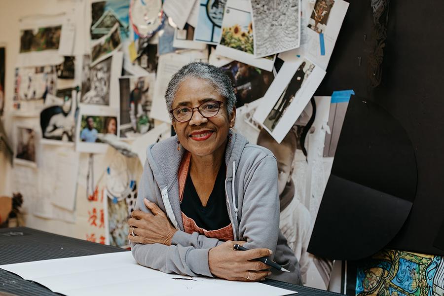 A woman sitting and leaning on her elbows at a desk with artworks taped on a wall behind her