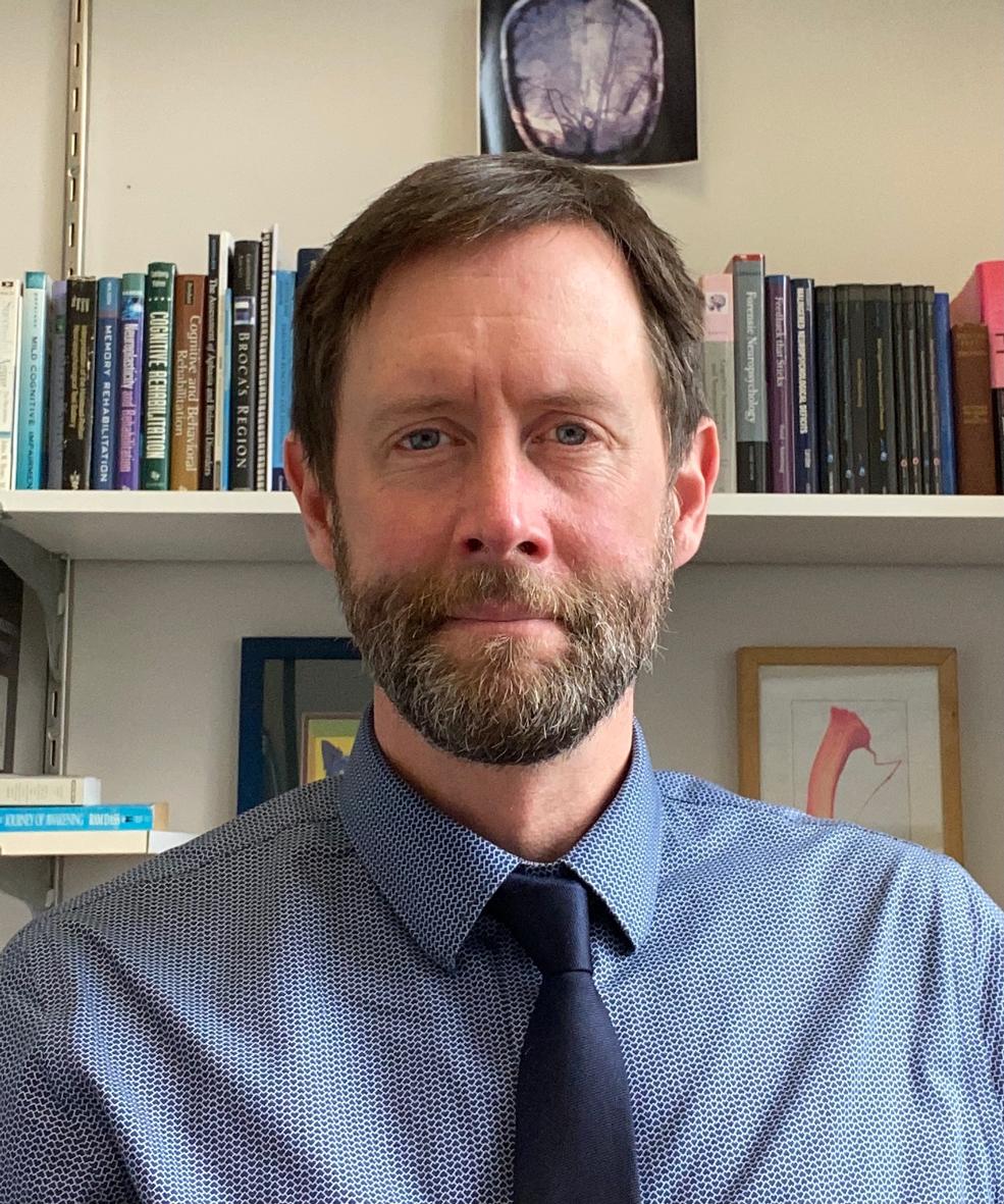 Photograph of Kris Rhoads, a man in a blue collared shirt with a blue tie, short brown hair and a beard and mustache, with a shelf of books and artwork on the wall behind him