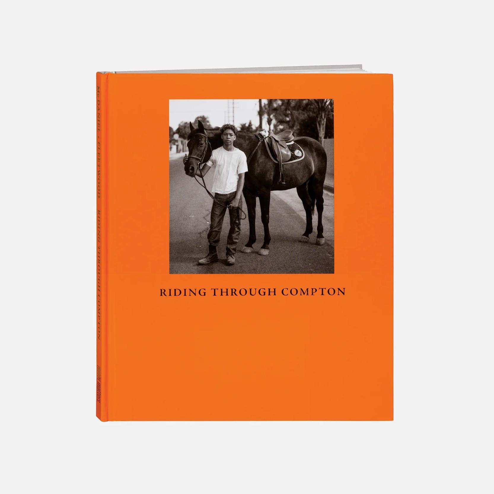 A book with an orange cover and a photo of a young black person standing with a horse with the words "Riding Through Compton" underneath