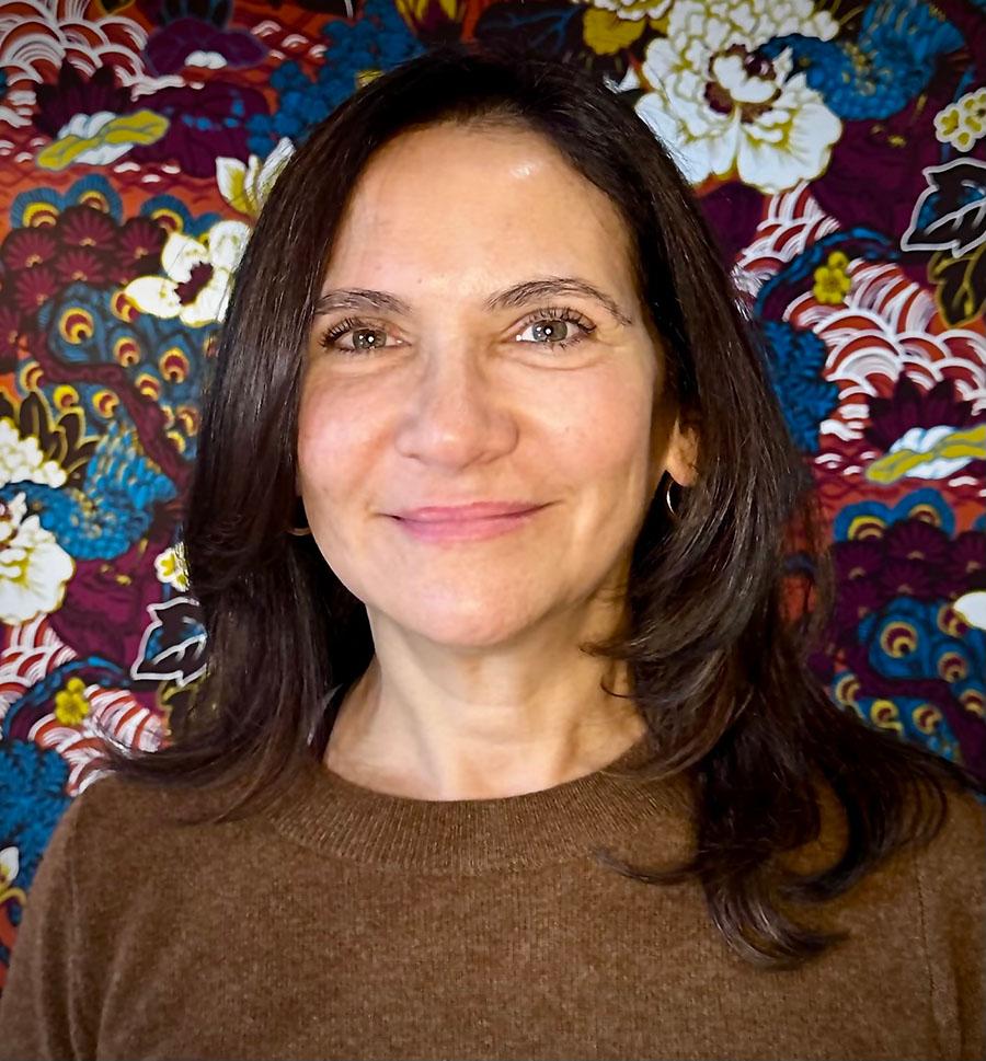 A slightly smiling person with brown hair in front of a bright, detailed, patterned fabric