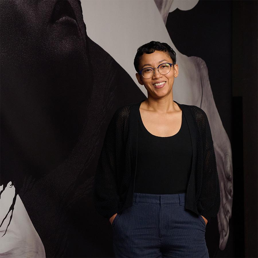 A person with short hair standing in front of a wall with a large artwork