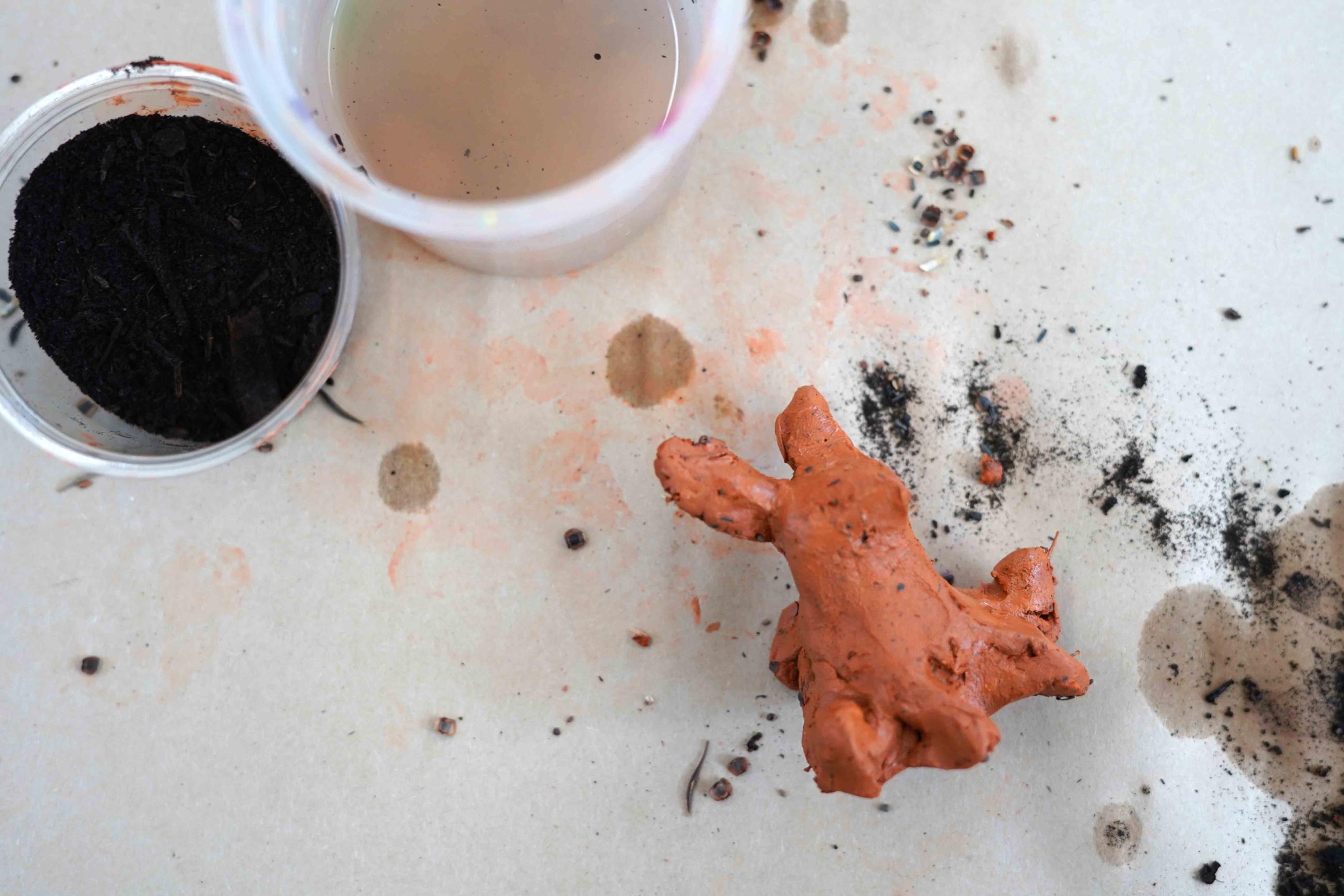 Photo of a cup with water, a cup with soil, and a small clay sculpture shaped like a bunny