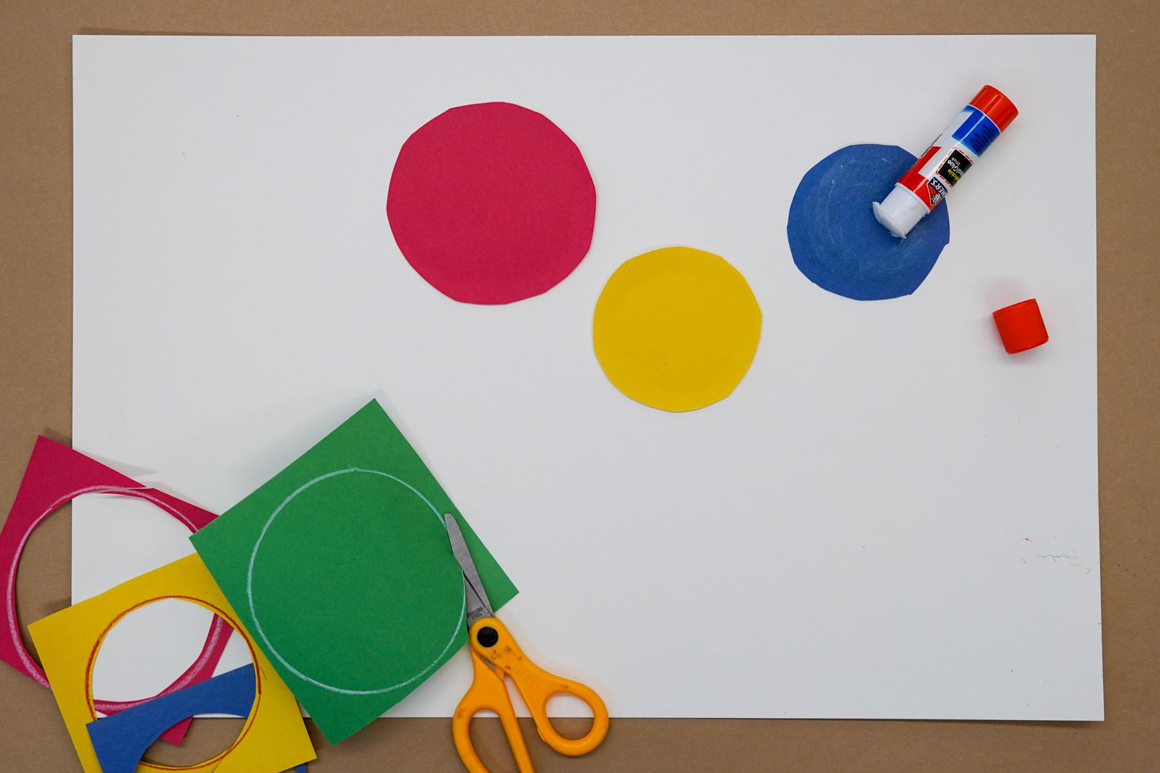 Photo of a white piece of paper with three colorful circles of construction paper on top along with a glue stick and scissors and paper remnants in the lower left corner