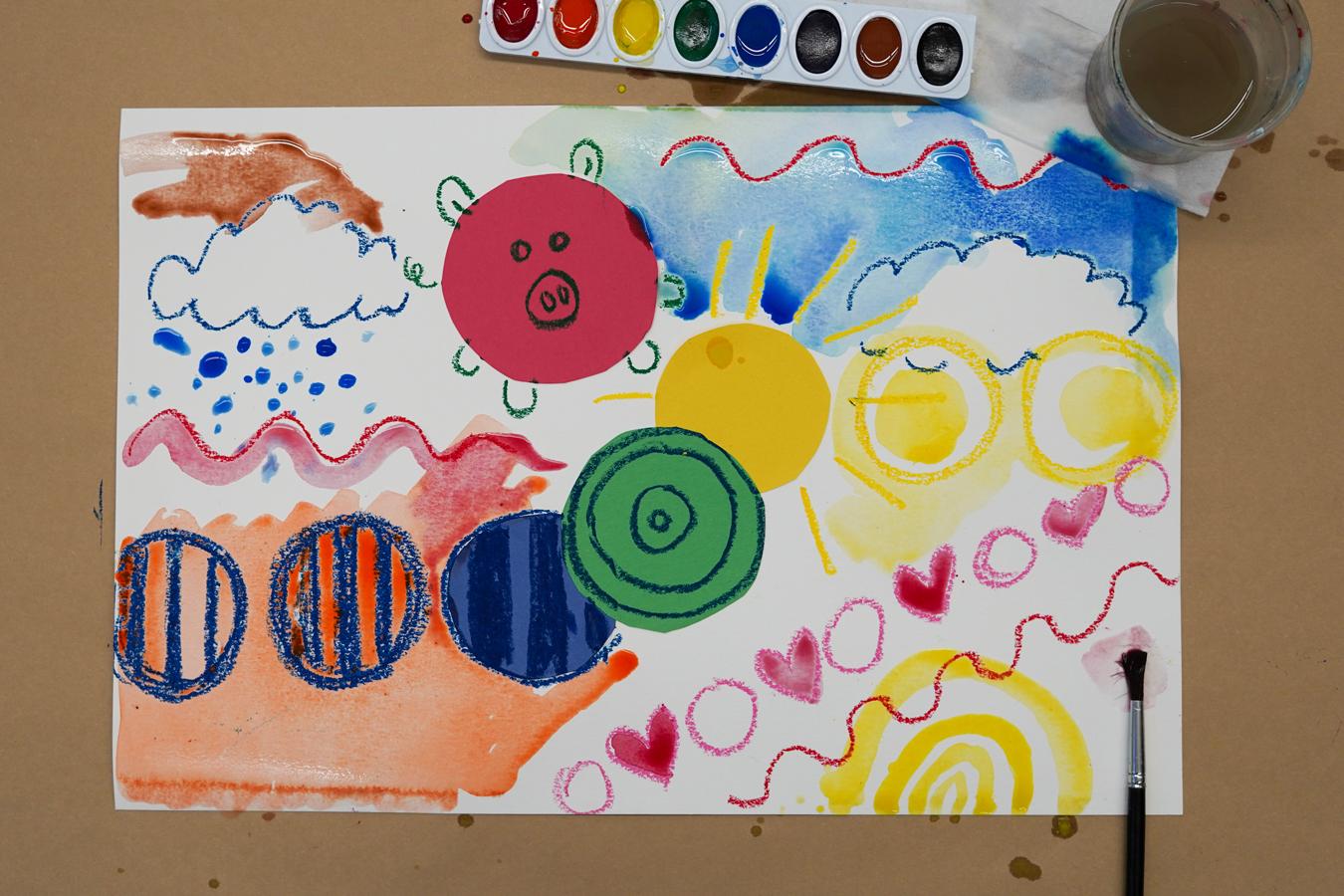 Photo of a piece of paper with colorful construction paper dots on it and colorful doodles as well as colorful watercolors; a set of watercolors, a brush, and a glass of water sit around the paper