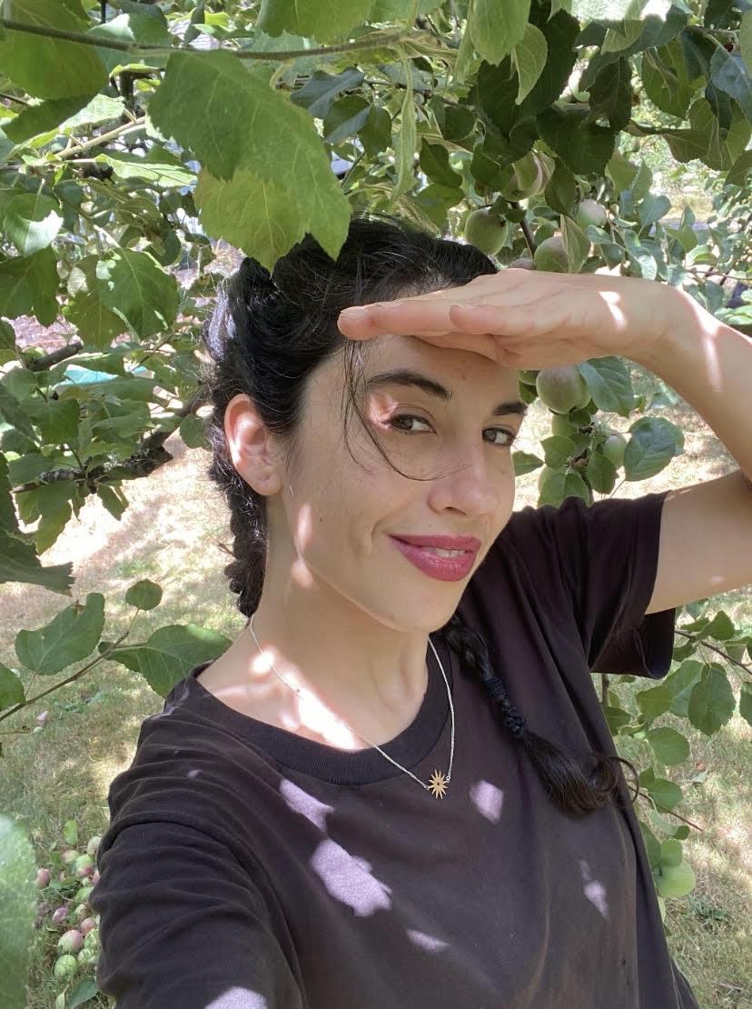 Photo of Ursula Brown, a woman with dark hair pulled back wearing a black t-shirt, surrounded by the leaves of a tree