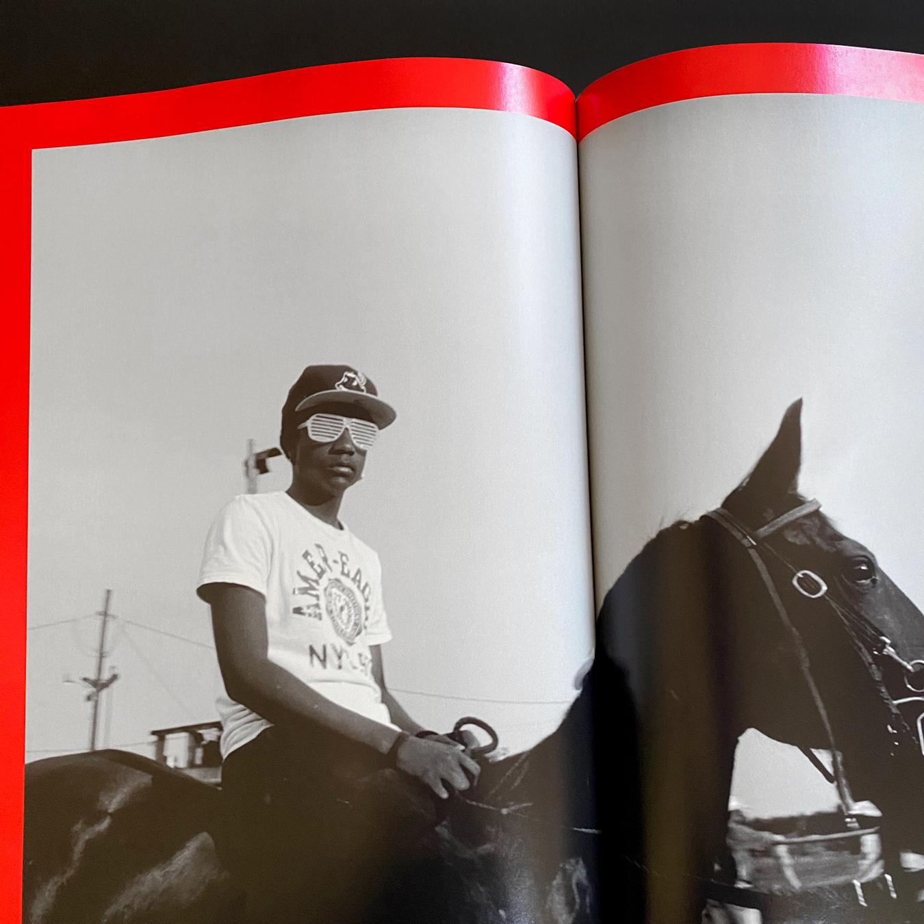 Photo of a young Black person wearing sunglasses and a baseball hat riding a horse, which spans two pages in an open book