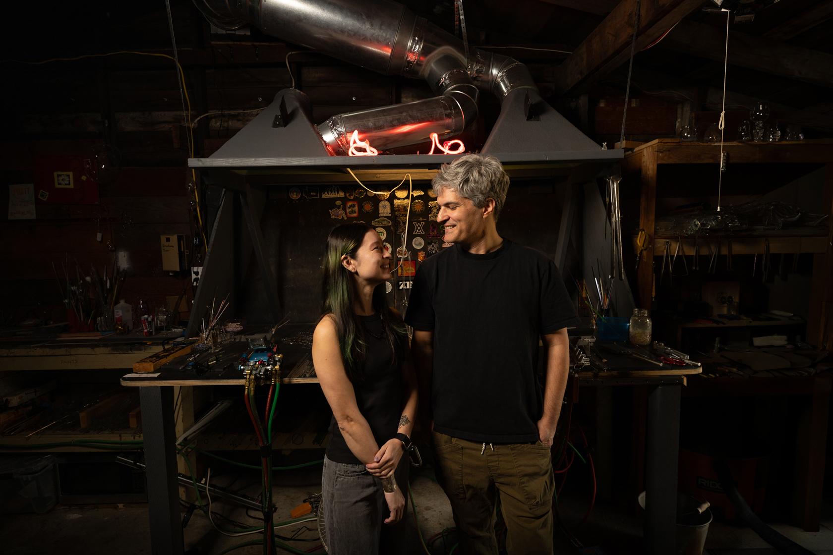 A couple standing in a flameworking glass studio smiling at each other