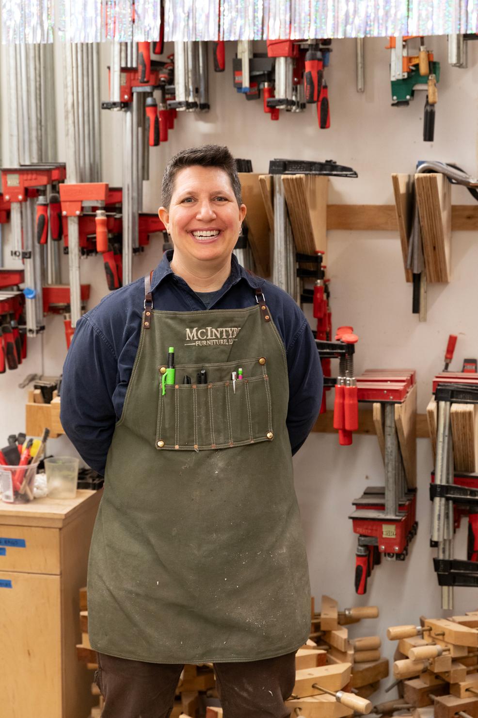 Kim McIntyre smiling and standing in a woodshop with an olive work apron on